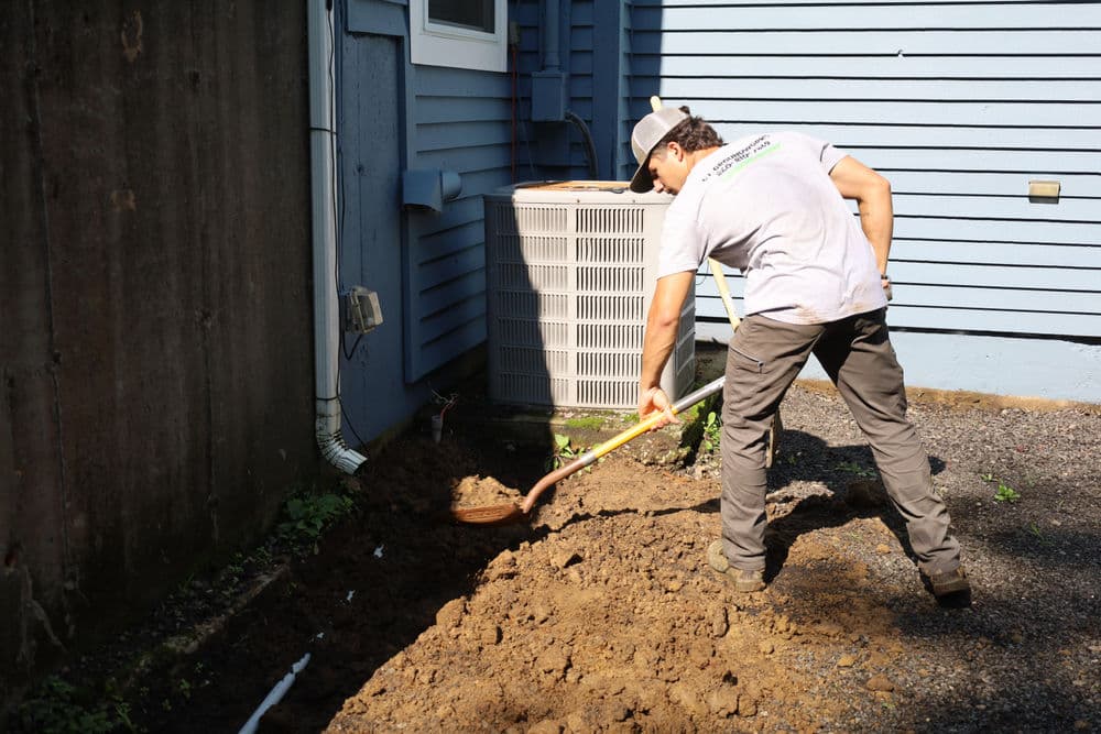 Person digging a trench in front of a house for landscaping or plumbing installation.