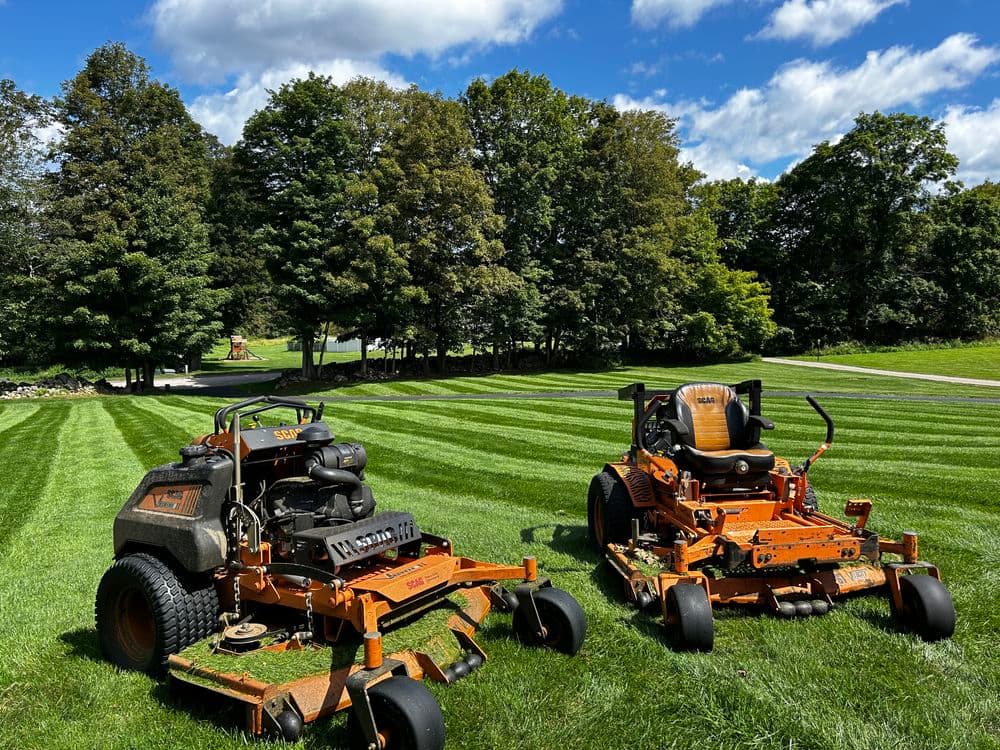 Two orange lawn mowers on freshly cut green grass under a blue sky with clouds.