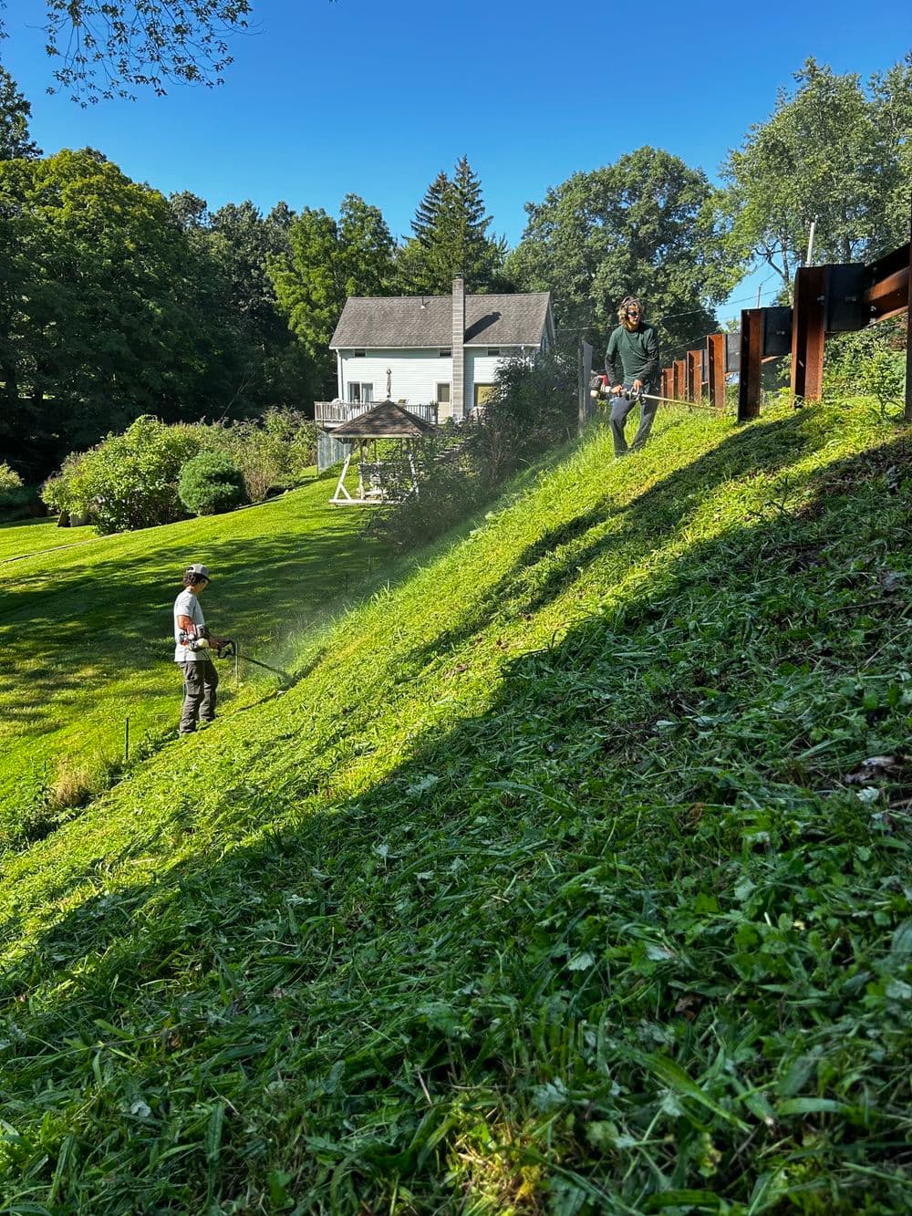 Two people mowing a sloped lawn near a house in a bright, sunny garden setting.
