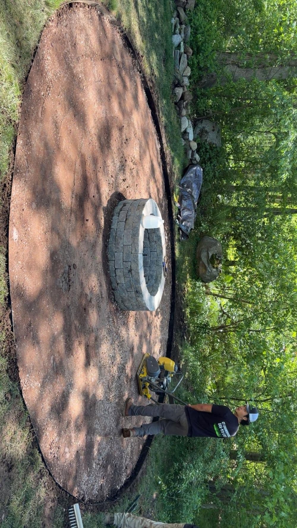 Stone fire pit installation in a grassy garden surrounded by trees and landscaping tools.